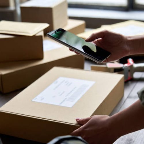 Female warehouse worker seller, small stock business owner holding phone scanning retail package parcel bar code on commercial shipping box delivery order on smartphone using mobile app.