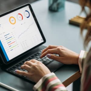 Woman in striped shirt working on tablet displaying colorful graphs and data analysis in a business environment.